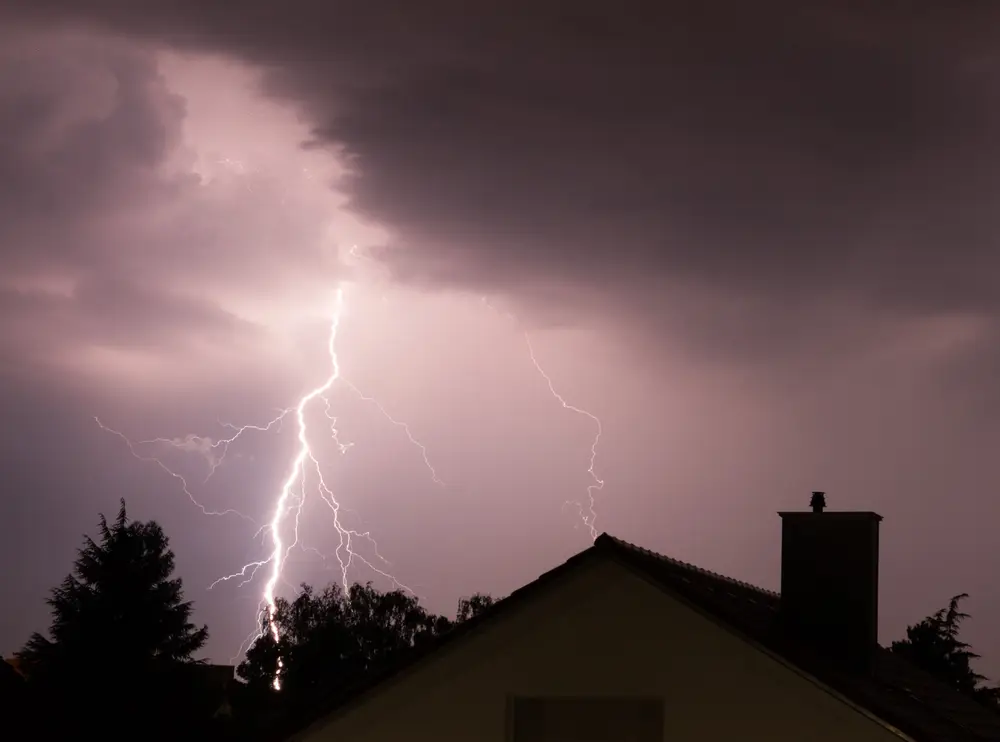 A bright lightning bolt streaks across a dark, cloudy sky above the silhouette of a house and trees at night, casting eerie shadows and highlighting where expert exterior contractors near me might work.