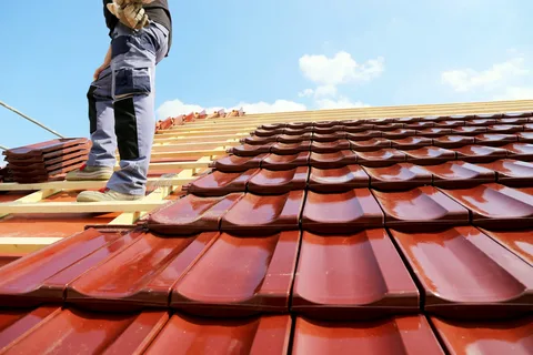 At a picturesque location, a worker places red roof tiles onto a slanted roof under a clear sky.