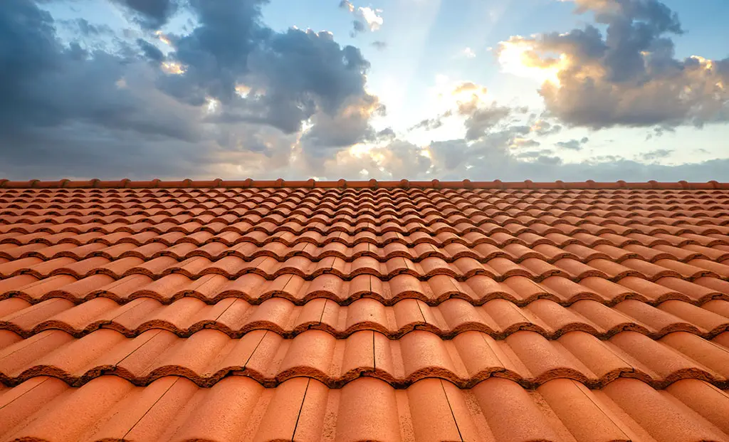 A close-up of a red clay tile roof in a picturesque location, with a partly cloudy sky and sunlight peeking through the clouds in the background.