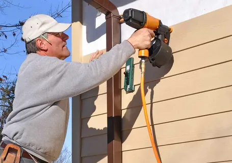 A person wearing a cap uses a nail gun to install siding on the exterior wall of a building at a construction site.